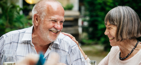 A 50-something husband and wife sit close to each other and share a laugh while dining outside on a warm day. 