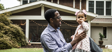 Sitting in front of a home, a father happily steadies his 1-year-old daughter as she stands.